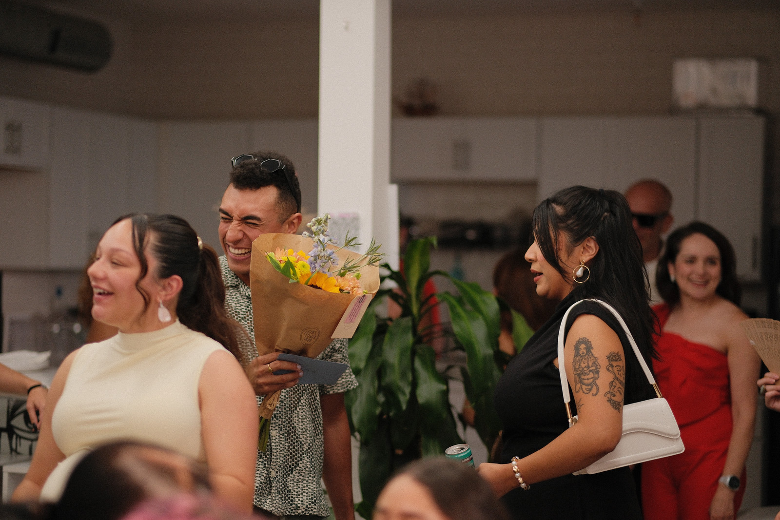 A guest laughs with his eyes shut while holding a wrapped bouquet of yellow and lavender flowers, surrounded by friends and family in the studio kitchen.