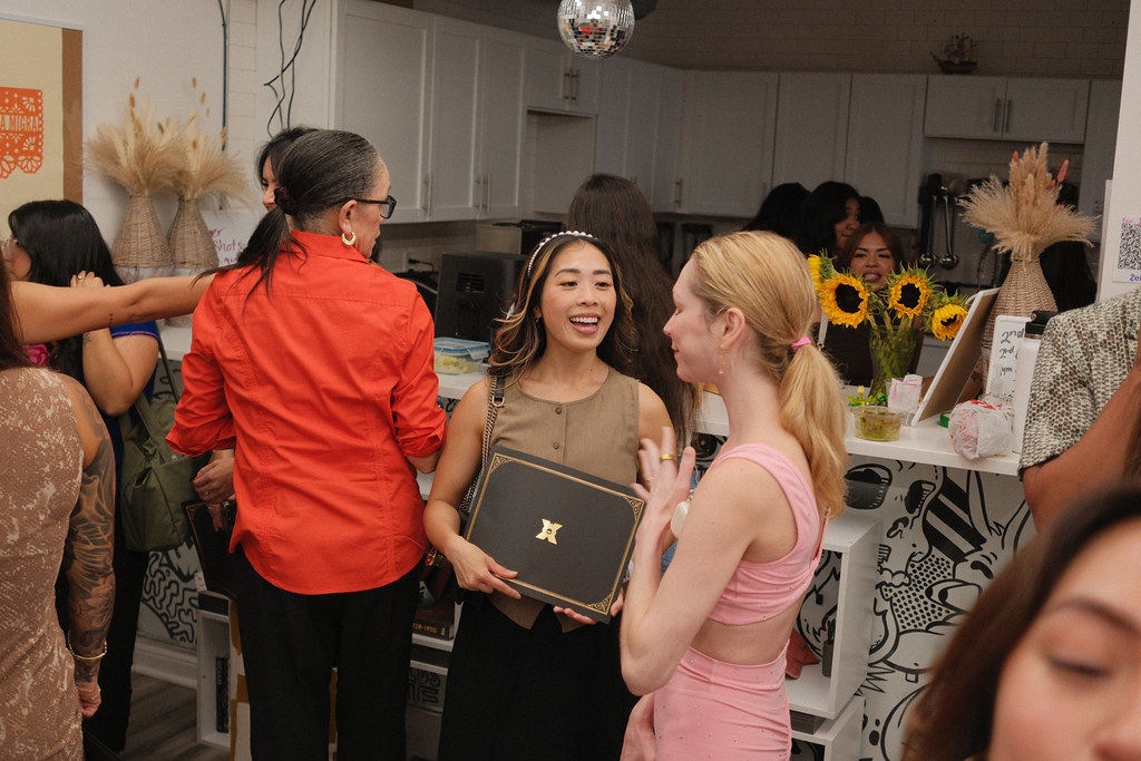 A graduate holds her black and gold LSP diploma folder against her chest, talking with two friends beside a counter of sunflowers, a disco ball hanging overhead.