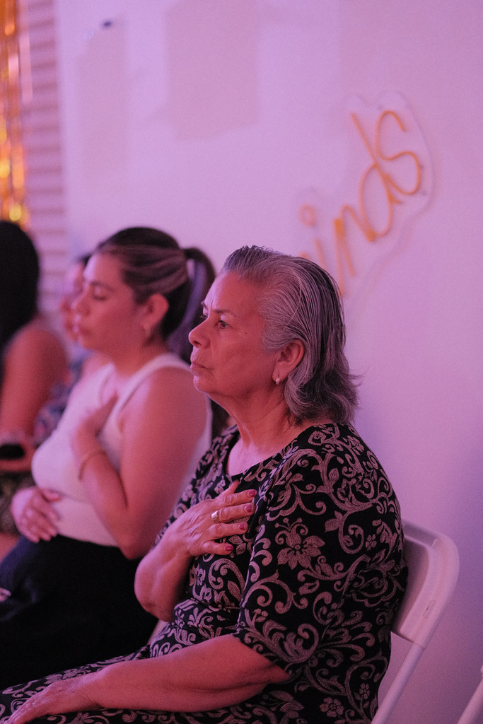 An older woman in a black and silver brocade dress sits with her hand over her heart, eyes forward, a younger woman doing the same behind her, both lit in warm pink light.