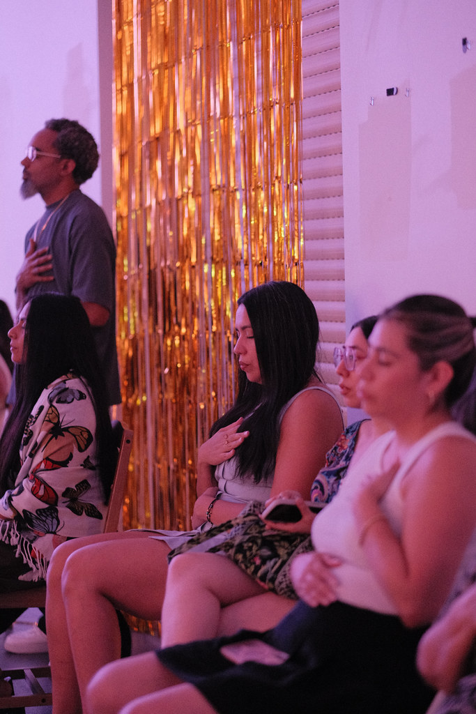A row of guests seated in front of a gold fringe curtain with their hands over their hearts and eyes closed, one man standing behind them doing the same.