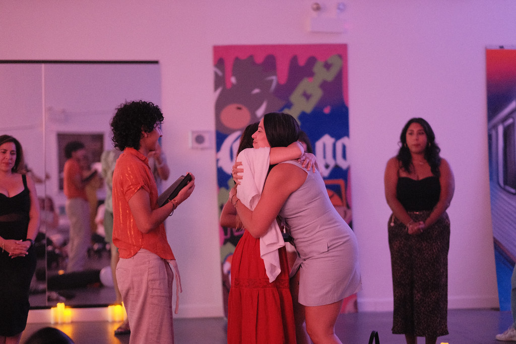 Two graduates hold each other in a long hug while a classmate waits alongside them holding diploma folders, candles glowing at the base of the mirror.