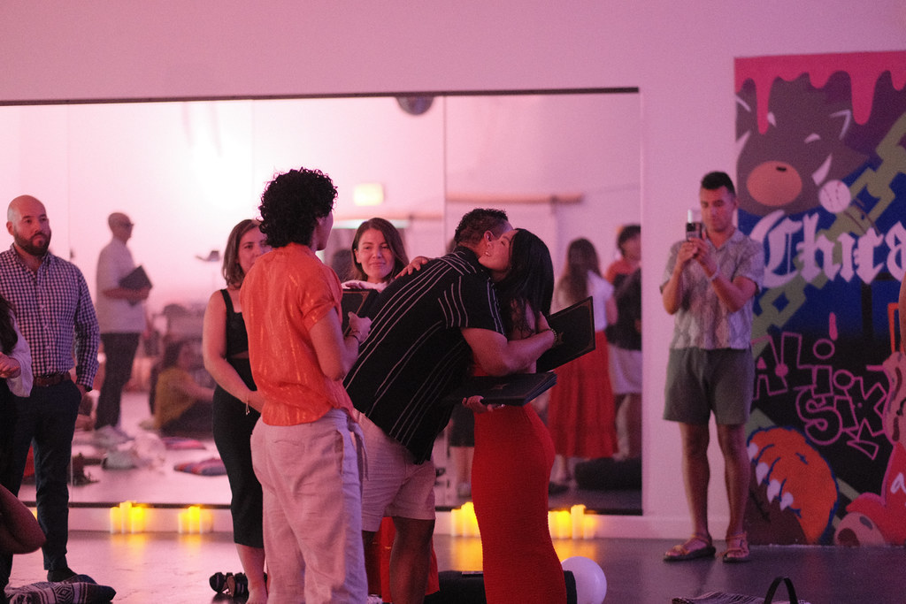 A graduate is lifted into a hug, her diploma folder still in her hand, while a guest films the moment on his phone from across the room.