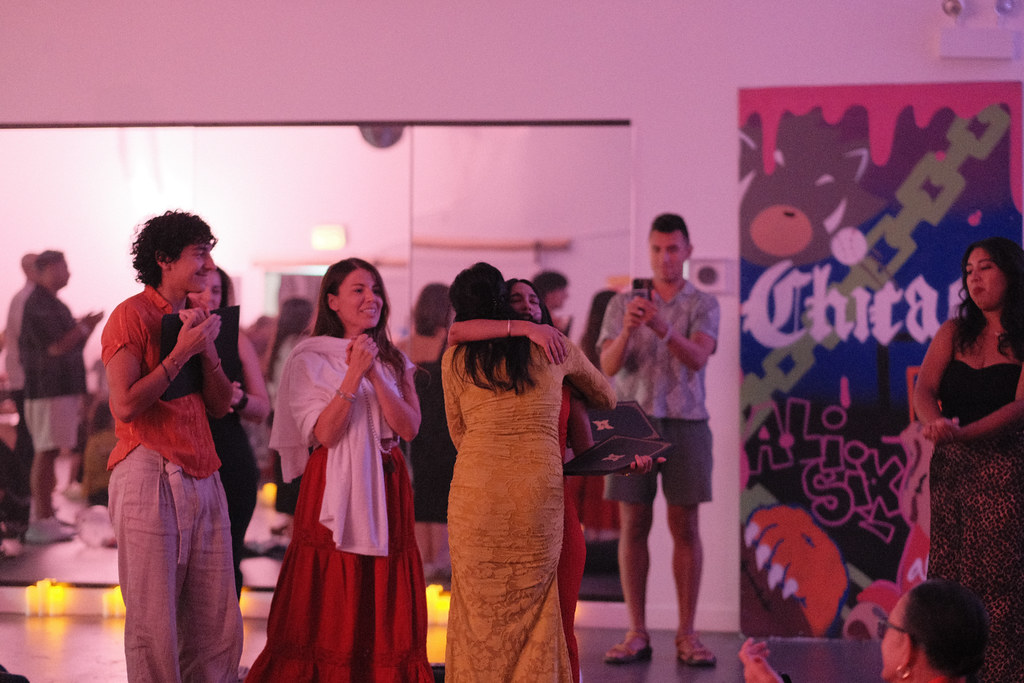 The room applauds as two graduates embrace, one in a gold dress holding her diploma folder behind her friend's back, rows of candles lining the floor.