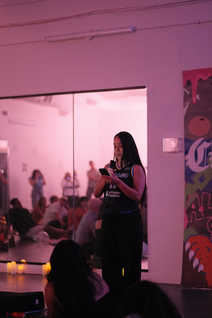 Vanessa Tirado speaks into a handheld microphone while reading from her phone, wearing a Chicago basketball jersey, the seated room reflected in the mirror behind her.