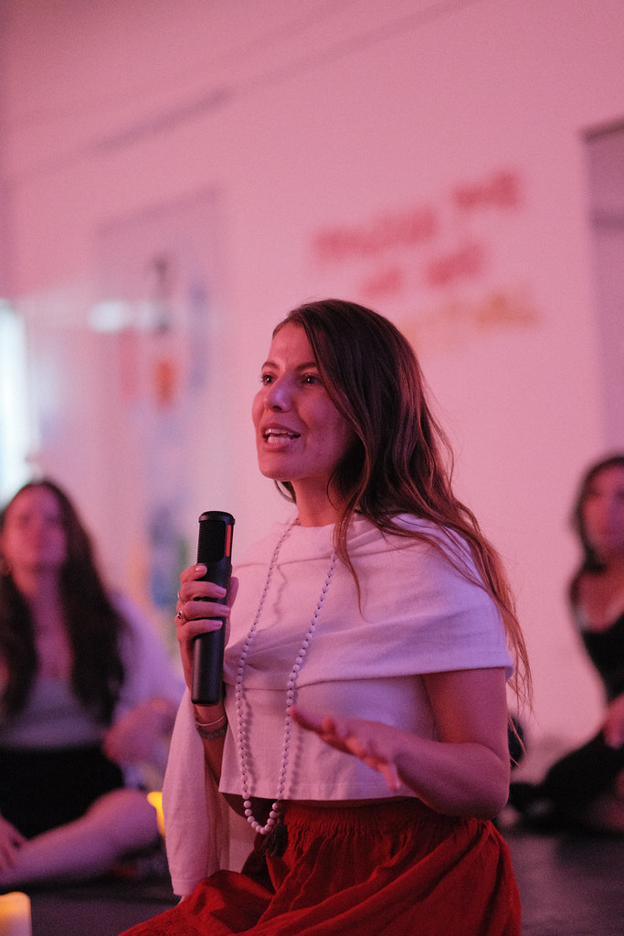 Margarita Quiñones-Peña speaks mid-sentence with a microphone, in a white top and long strand of pearls, graduates seated on the floor around her.