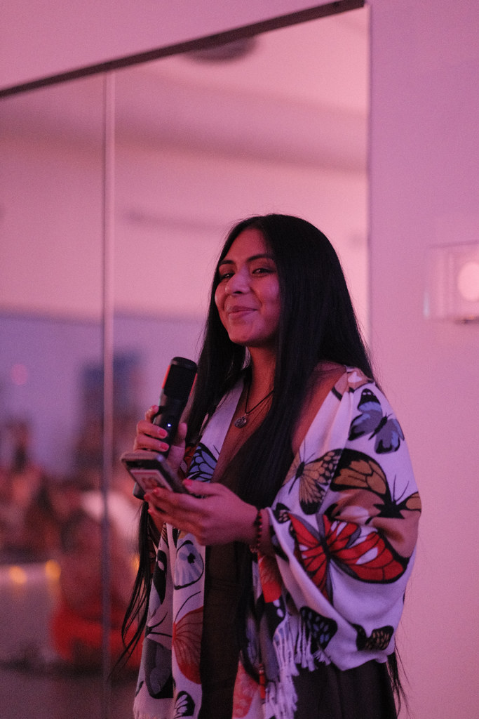 Yesi Peyret smiles as she speaks into a microphone, holding her phone in her other hand, wearing a cream rebozo printed with monarch butterflies.