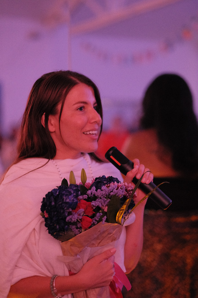 Margarita Quiñones-Peña laughs mid-speech with the microphone in one hand and a bouquet of blue hydrangeas and roses held against her chest.
