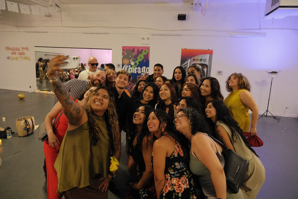 A crowd of graduates and guests packs into a group selfie, one arm stretched out holding the phone, everyone leaning in and laughing.