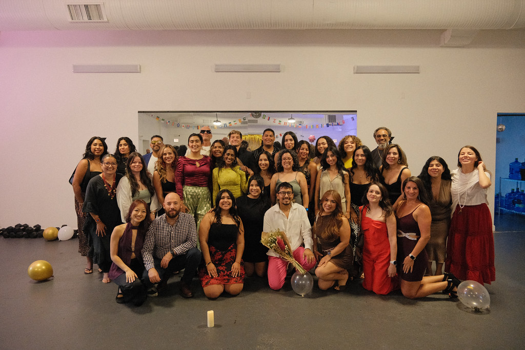 The full graduating group poses together in warm pink light, some kneeling in front, gold and white balloons on the studio floor around them.
