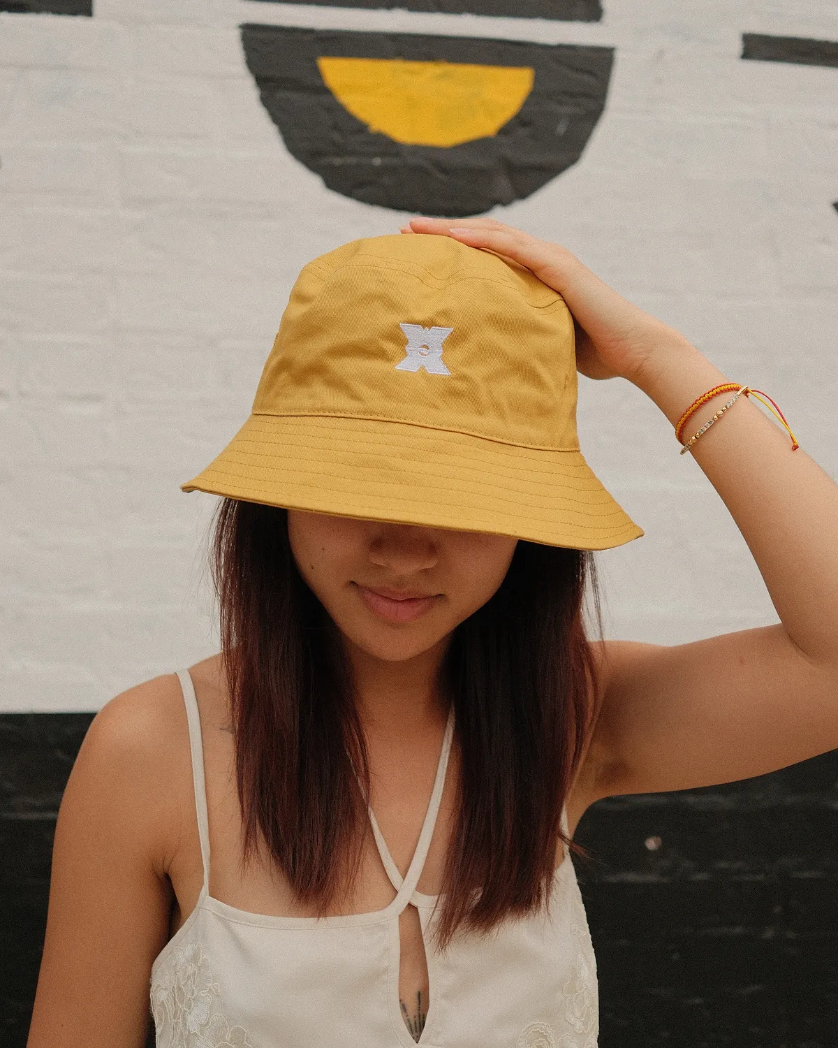 Mustard bucket hat with the embroidered LSP X, worn in front of the X mural