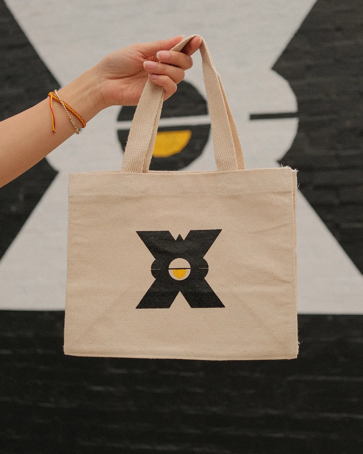 Natural canvas tote with the large LSP X and sun logo, held up in front of the X mural